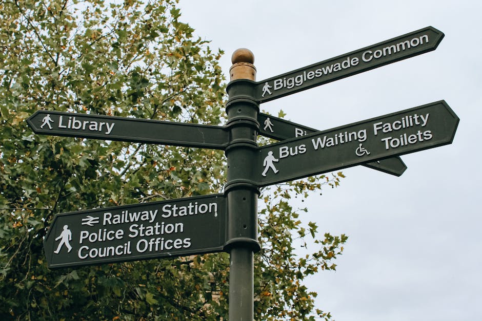 A black directional signpost with multiple arrows pointing towards local amenities such as Biggleswade Common, Library, Bus Waiting Facility, Toilets, Railway Station, Police Station, and Council Offices, situated outdoors against a background of leafy trees and an overcast sky. The sign features white text and icons indicating pedestrian access, with some arrows including wheelchair accessibility symbols. The scene is well-lit by natural daylight, highlighting the clean and clear appearance of the sign. This type of surface cleaning and maintenance by [COMPANY_NAME] ensures signage remains visible and hygienic for the community, aligning with hygienic standards and general urban upkeep recommended for public spaces.