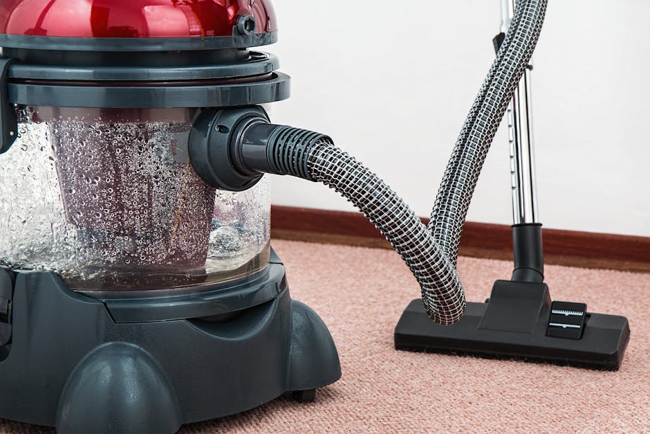 A close-up view of a commercial carpet cleaning vacuum with a transparent canister filled with water and foam. The vacuum features a flexible, striped hose connected to a black floor attachment, positioned on a pinkish-beige carpet in a well-lit indoor setting with a white wall and wooden skirting board in the background. The scene highlights surface cleaning and deep carpet sanitisation, as performed by professionals like Cleaners SW4, to ensure thorough hygiene and maintenance of residential or commercial flooring surfaces.