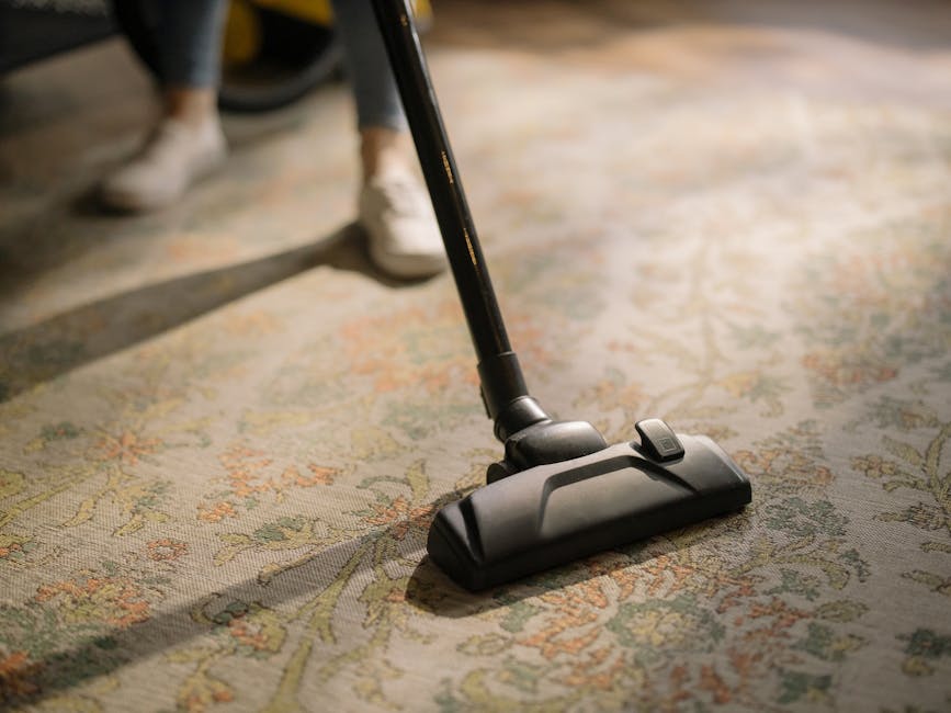 Close-up image of a person vacuuming a carpet in a residential setting, with a black vacuum cleaner attachment on an ornate, patterned carpet featuring floral motifs in shades of beige, green, and orange. The room is lit by natural daylight, creating shadows on the carpet surface. The person's legs are visible in the background, wearing dark pants and white shoes, and the vacuum cleaner hose is positioned centrally, emphasizing surface cleaning and deep hygiene maintenance, as offered by Cleaners SW4 for domestic and commercial properties near Clapham Common SW4.