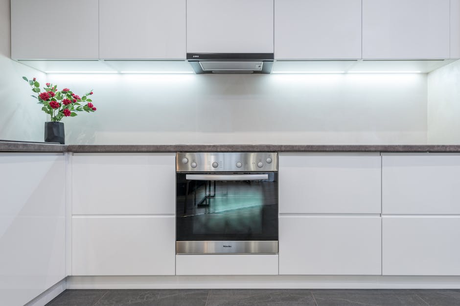 A modern kitchen with a sleek, white minimalist design featuring smooth, handle-less cabinets and a grey countertop. The stainless steel oven with a glass door is built into the cabinetry, and a black range hood is mounted above it. The countertop has a black vase containing a bouquet of pink and red roses with green leaves, adding a touch of color. The room is well-lit with bright, even lighting, reflecting off the clean, polished surfaces, indicating a high level of surface cleaning and maintenance by Cleaners SW4. The overall cleanliness and shiny finishes suggest recent deep cleaning and sanitisation, supporting professional domestic or commercial cleaning practices aligned with surface and kitchen deep cleaning standards.