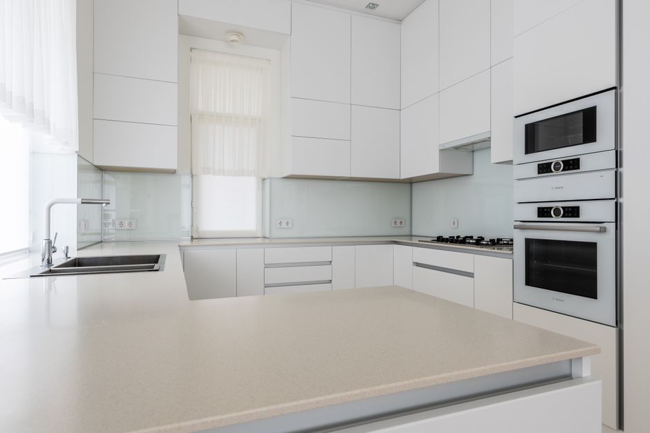 A modern, minimalist kitchen with white cabinetry and a light-colored solid surface countertop. The room features a stainless steel sink with a chrome faucet situated beneath a window that allows natural light to fill the space. The kitchen includes built-in stainless steel appliances, such as a microwave, oven, and a gas cooktop, all integrated into the cabinetry. The backsplash is a clear, glossy glass panel that reflects the cleanliness of the surfaces, with no visible dirt or stains. The entire area appears freshly cleaned and well-maintained, with smooth, dust-free surfaces and pristine appliances, exemplifying a professional level of surface cleaning and sanitisation in domestic kitchen spaces, as provided by Cleaners SW4.