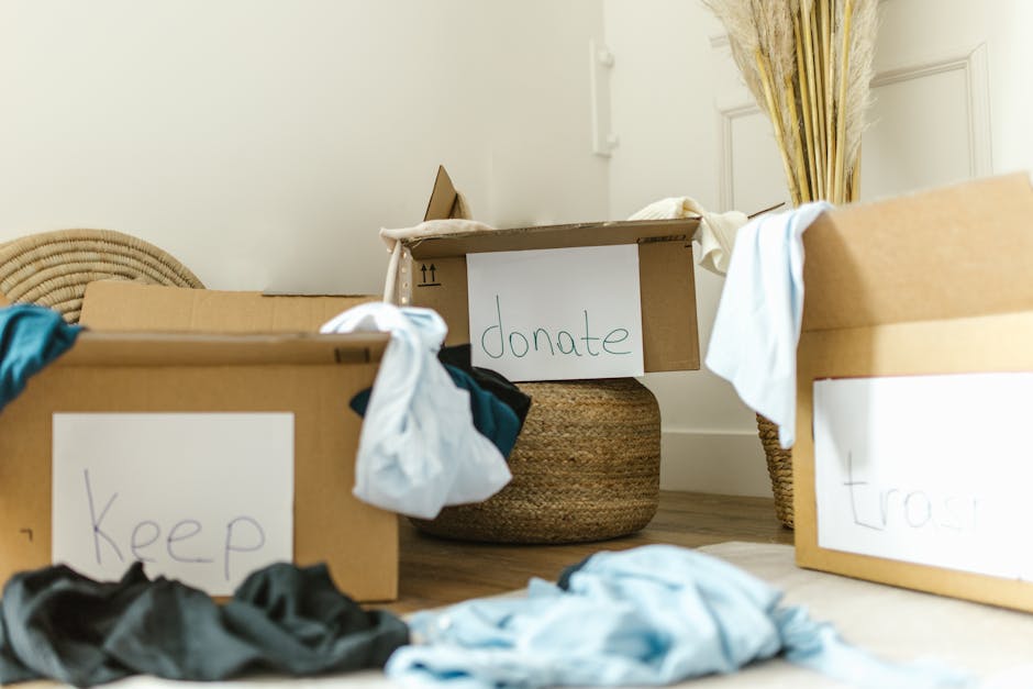 Indoor scene showing cardboard boxes with handwritten labels such as 'keep', 'donate', and 'trash' placed on a wooden floor. The boxes contain various textiles and clothes, with some draped over the e