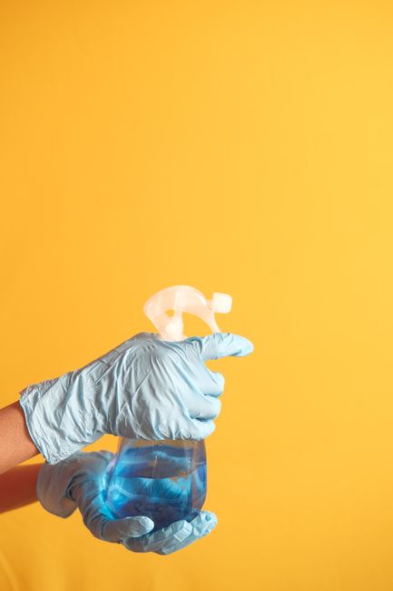 A hand wearing a light blue disposable glove holding a clear spray bottle filled with blue cleaning solution, with a white spray nozzle, against a solid yellow background. The image highlights surface cleaning and sanitisation processes associated with domestic or commercial cleaning, as provided by Cleaners SW4, emphasizing hygiene and deep cleaning services available for properties in Clapham Junction SW4. No additional objects or surfaces are visible in the image, focusing solely on the cleaning product and hand.