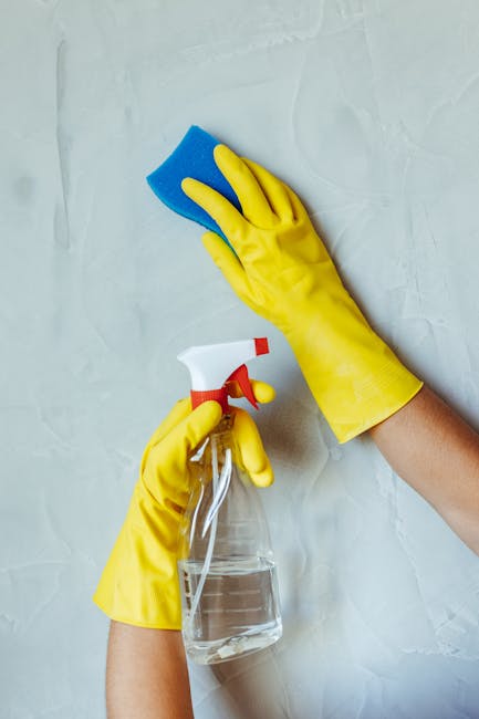 A hand wearing a light blue disposable glove holding a clear spray bottle filled with blue cleaning solution, with a white spray nozzle, against a solid yellow background. The image highlights surface cleaning and sanitisation processes associated with domestic or commercial cleaning, as provided by Cleaners SW4, emphasizing hygiene and deep cleaning services available for properties in Clapham Junction SW4. No additional objects or surfaces are visible in the image, focusing solely on the cleaning product and hand.