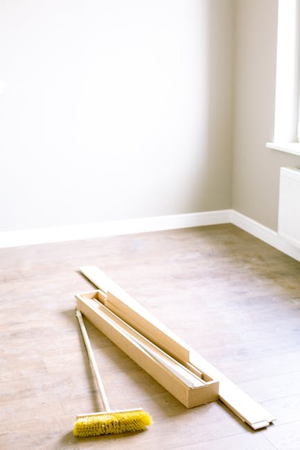 A clean, empty room with light wooden flooring and white walls illuminated by natural light from a window on the right. On the floor, there is a wooden window frame with glass panes laid flat, next to a long, yellow-handled dusting brush. The room appears freshly vacuumed and dusted, with no visible clutter or dust, showcasing proper surface cleaning and maintenance in preparation for end of tenancy, as part of a deep cleaning process by Cleaners SW4. The overall atmosphere is bright and tidy, emphasizing cleanliness and readiness for new occupancy.